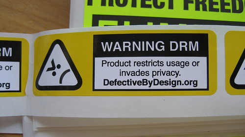 Um rolo de etiquetas autocolantes amarelos da campanha DefectiveByDesign.org.
Inclui o texto "Warning DRM: Product restricts usage or invades privacy.
DefectiveByDesign.org". À esquerda do está o logótipo da campanha, um
triângulo de cantos arredondados com uma representação em ASCII de uma cara
zangada >:(
